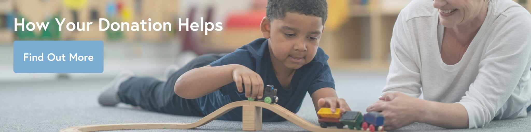 Young boy with dark hair laid on stomach playing with wooden trains, an adult helper sits nearby.