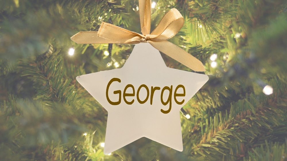 Goldd star shaped tree ornament with the name George in gold, hanging in front of an out of focus Christmas tree.