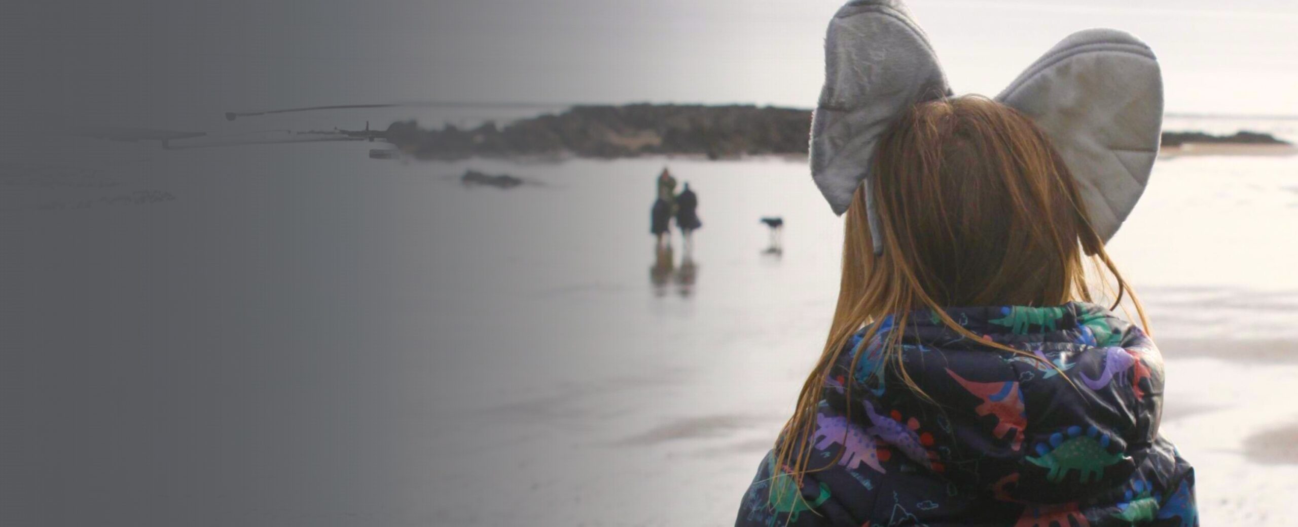 Photo with greay faded graident overlay. Young girl is standing with ehr back to camera, wearing elephant ears and looking out to sea.