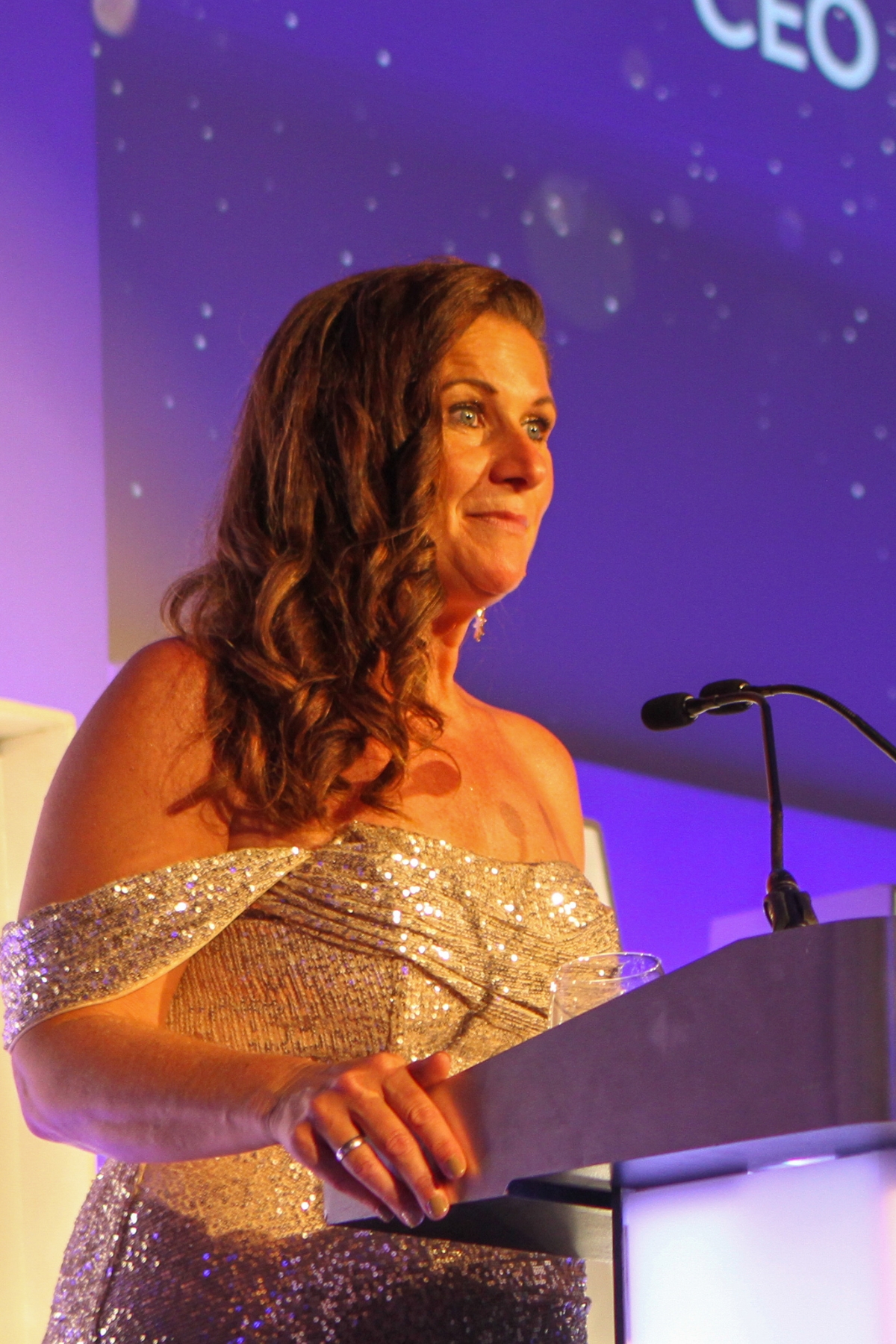Rhian Mannings MBE pictured standing in front of a podium with a microphone wearing a gold sequin evening gown