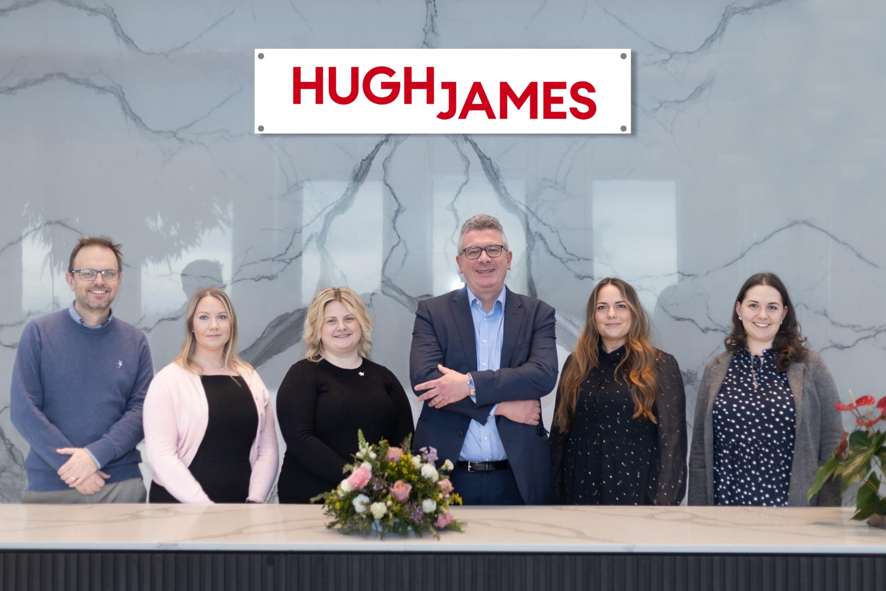 group photo of six individuals standing in front of marble wall with hugh james logo above them